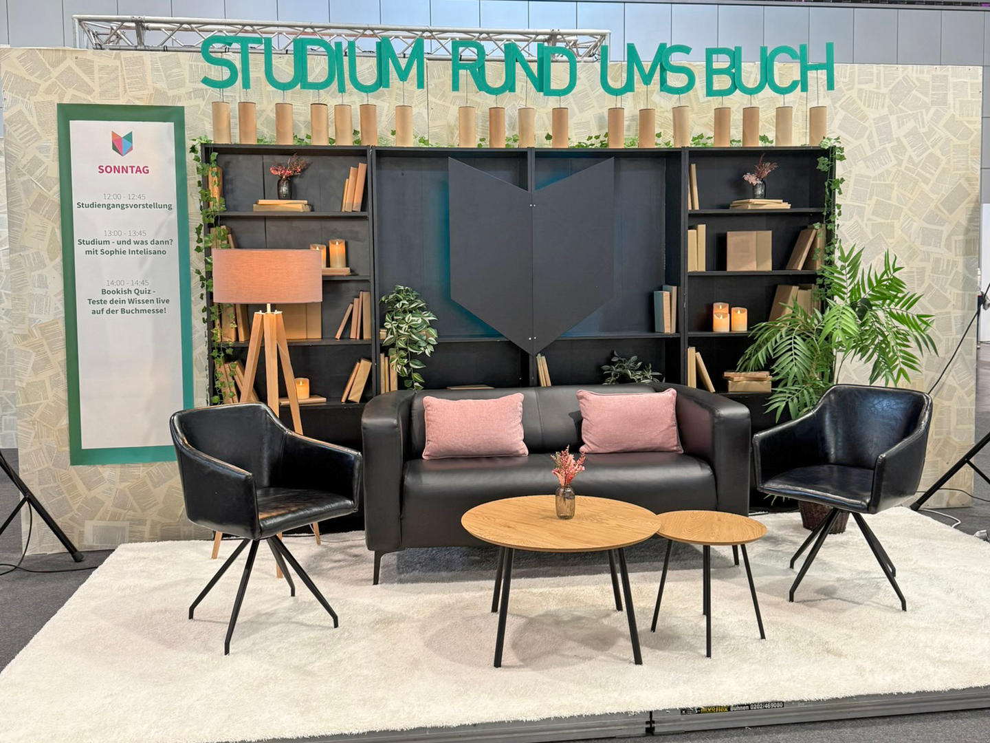 Buchmessestand mit einem Podium auf dem sich Sitzmobiliar und ein REgal befindet. Auf dem Regal stehen große Buchstaben, die die Worte "Studium rund ums Buch" ergeben