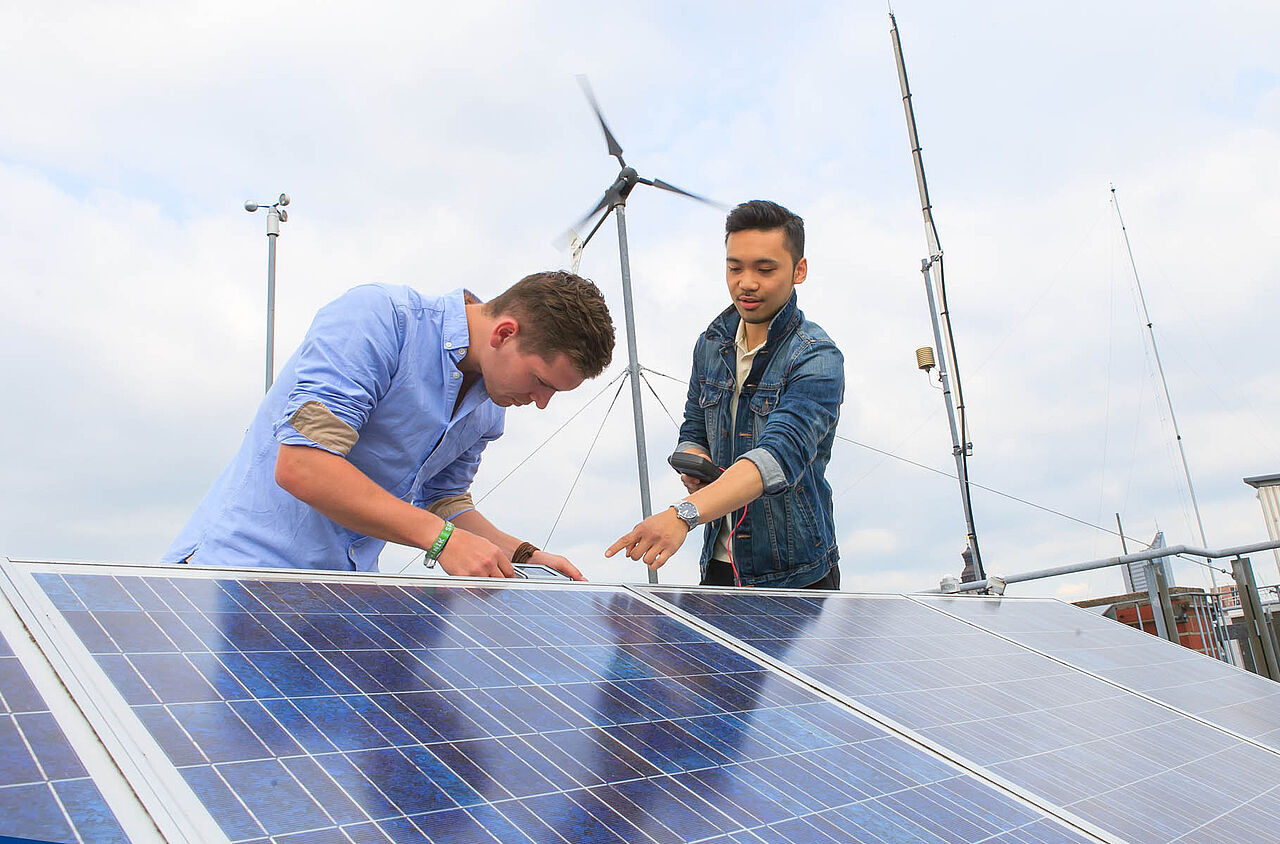 Zwei Studenten untersuchen Photovoltaikanlage. Im Hintergrund befindet sich eine Wettermessstation.