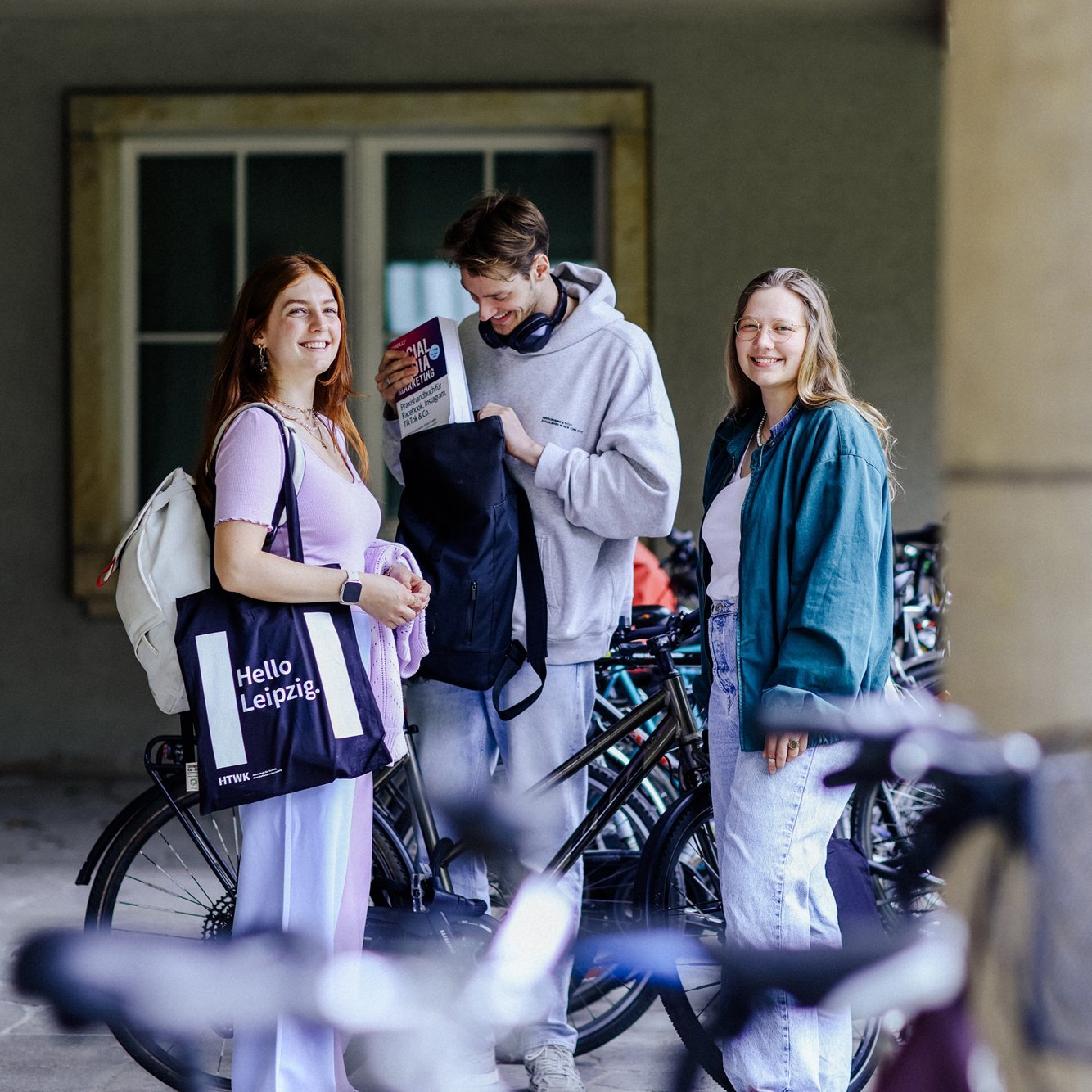 Drei Studierende stehen an einem Fahrradständer und lächeln in die Kamera.