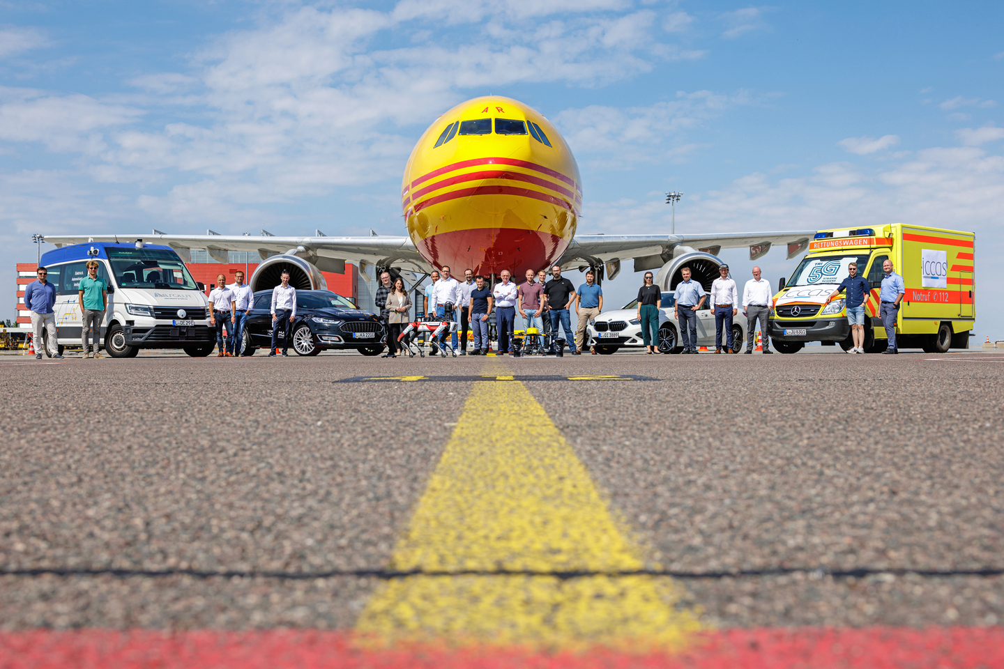 Verschiedene Transporter und Autos nebeneinander vor einem Frachtflugzeug aufgereiht, dazwischen Personen, die in die Kamera lächeln. 