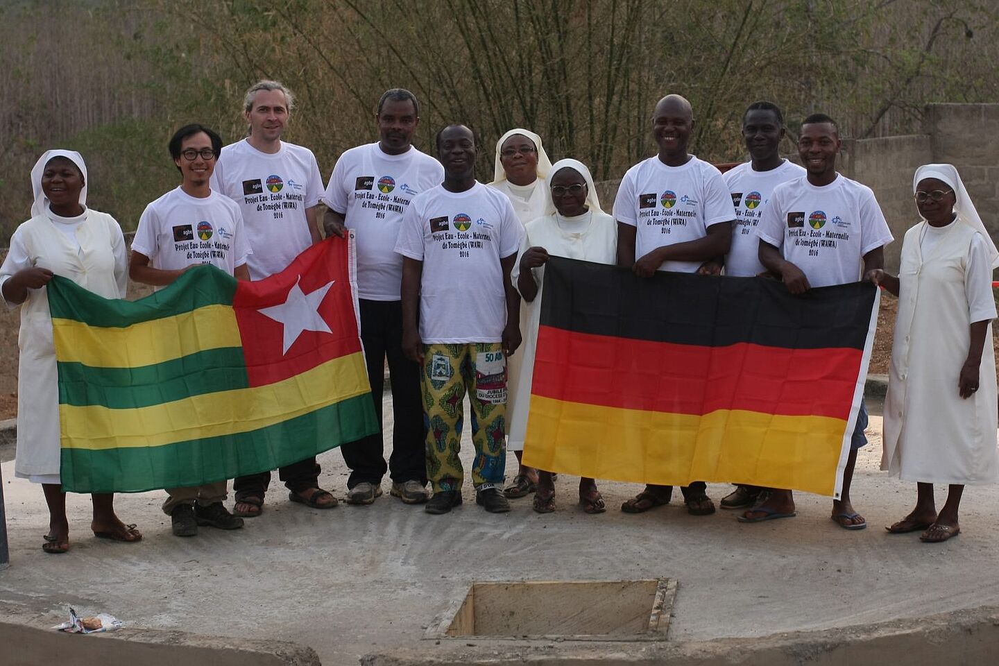 Gemeinsame Bauherren und zukünftige Träger Gruppenfoto mit Fahnen beider Nationen (Togo und Deutschland). Zu sehen sind knapp ein Dutzend Menschen nebeneinander, darunter deutsche Planer, einheimische Arbeiter und Ordensschwestern.
