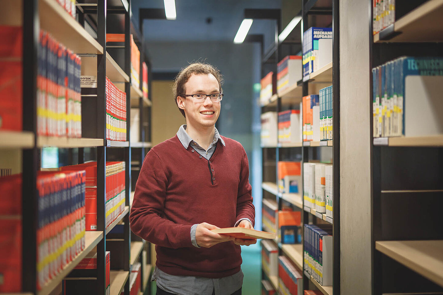 Ein Student zwischen zwei Bücherregalen in der Bibliothek hält ein Buch in der Hand.