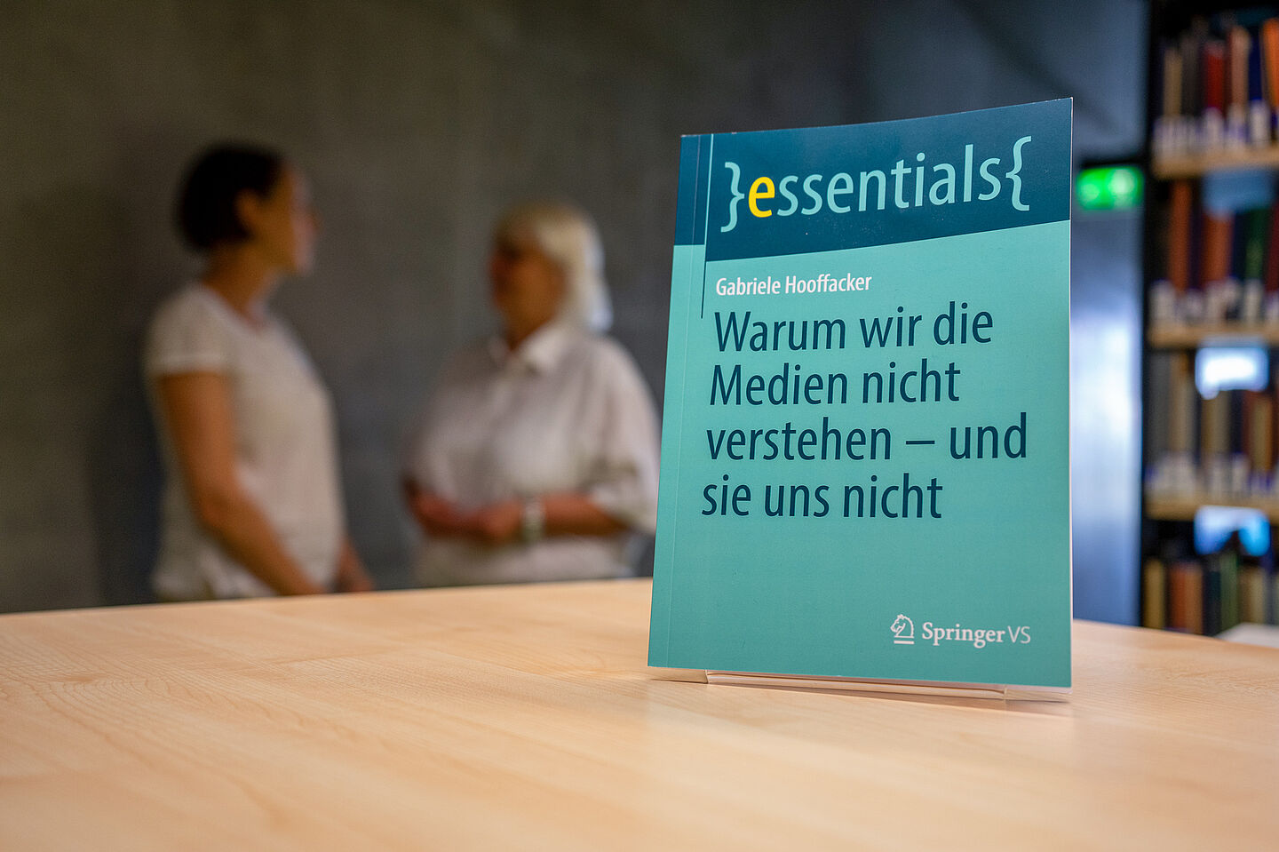 Im Vordergrund steht das Buch auf einem Tisch. Im Hintergrund sieht man beide Frauen im Interview.