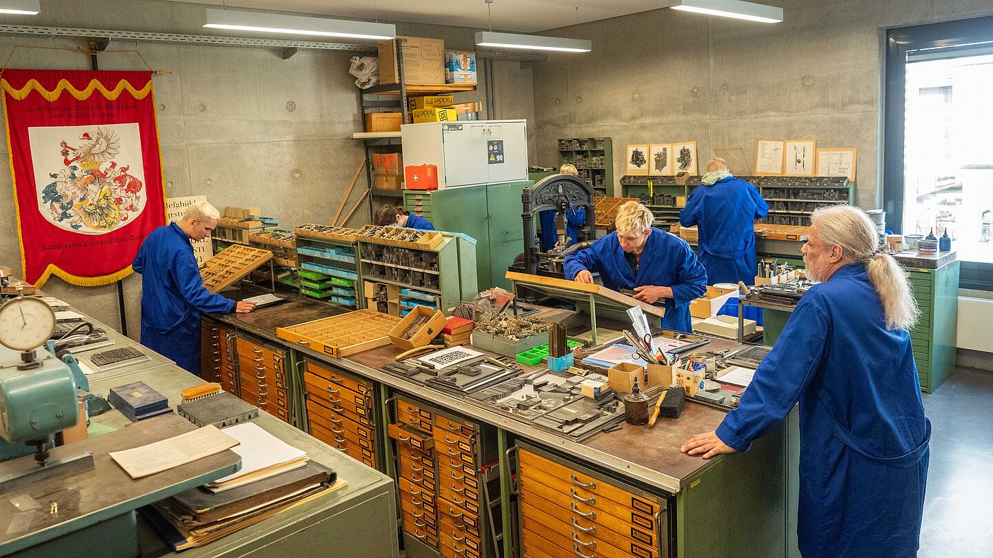 Blick in das Bleisatzlabor im Medienzentrum der HTWK Leipzig, in dem das Bleisatzpraktikum unter der Leitung von Prof. Dr. Michael Reiche stattfindet. Zusehen sind bei praktischer Tätigkeit zwei Studenten an großen Arbeitstischen mit Setzkästen und Prof. Dr. Michael Reiche, der diese beobachtet.