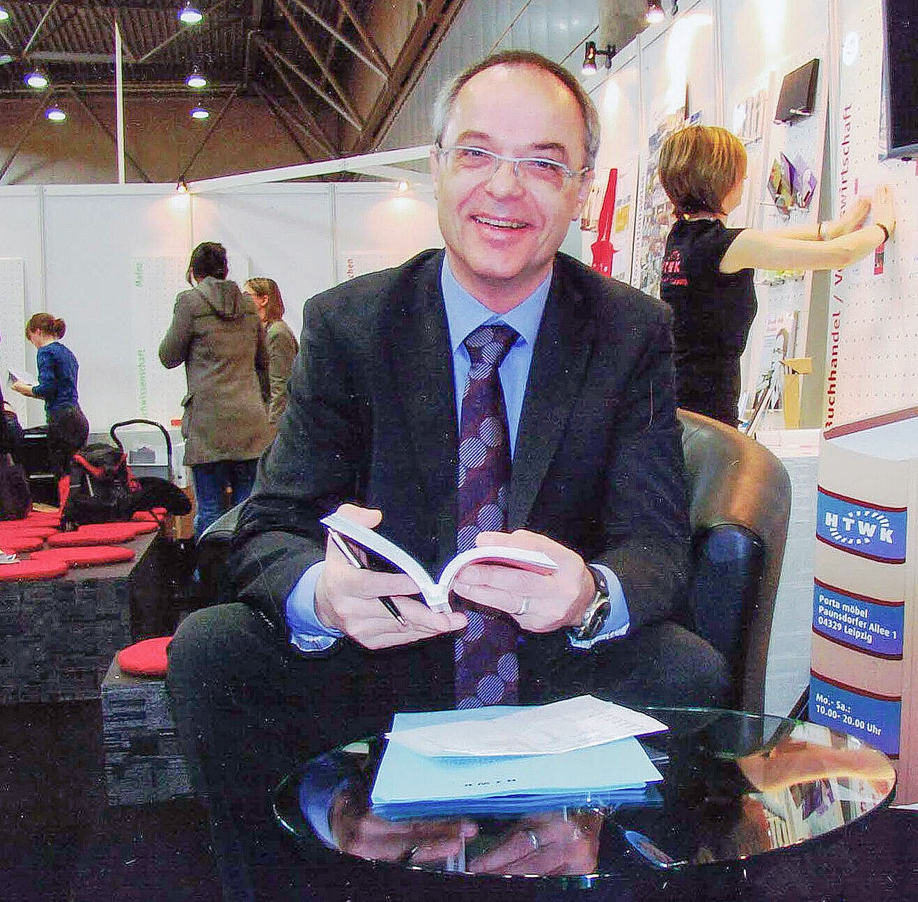 Professor sitzt im Vordergrund mit mehreren Büchern vor sich auf dem Tisch, im Hintergrund Messebesucher.