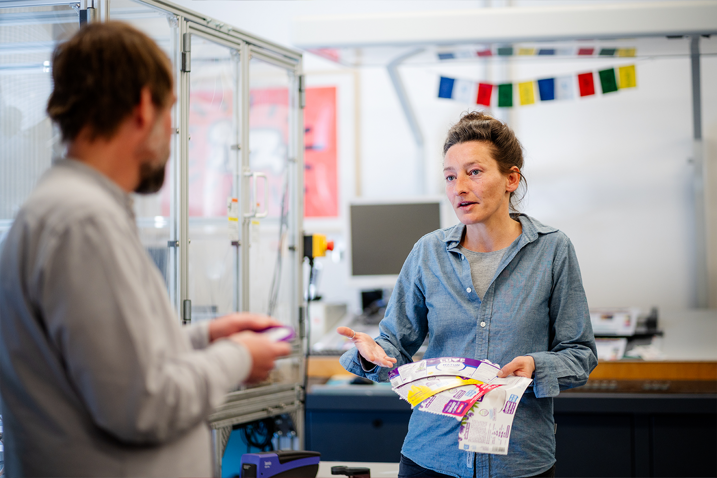 Prof. Dr. Ingo Reinhold und Katharina Roeber in einem Raum mit 3D-Druckmaschinen sprechen zueinander und halten beschichtete Testdrucke in den Händen.