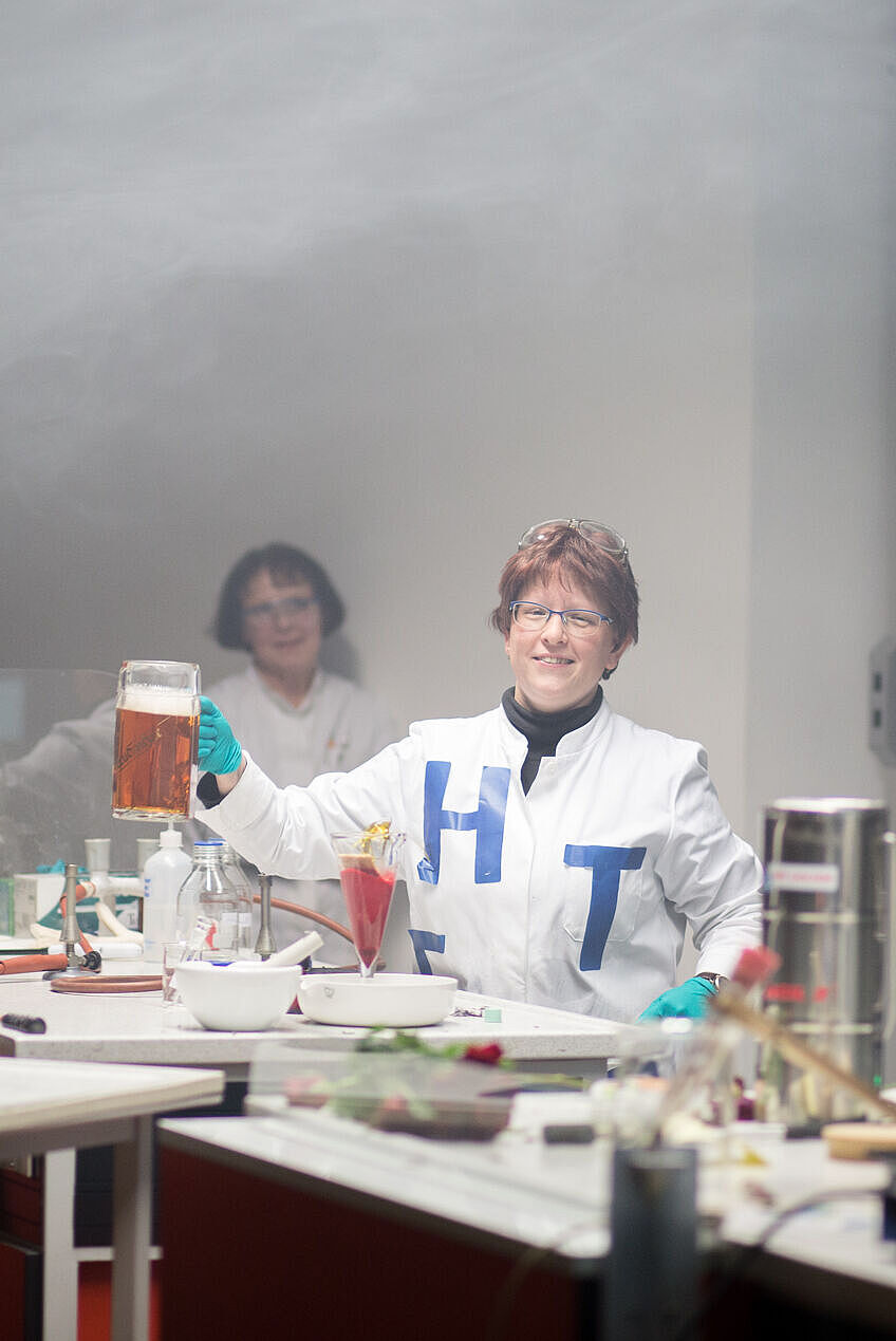 Andrea Berlich steht hinter dem Pult und hält in ihrer rechten Hand ein großes Bierglas, in dem ein chemischer Bierersatz enthalten ist. Über ihrem Kopf hängt der weiße Nebel vorangegangener Experimente.