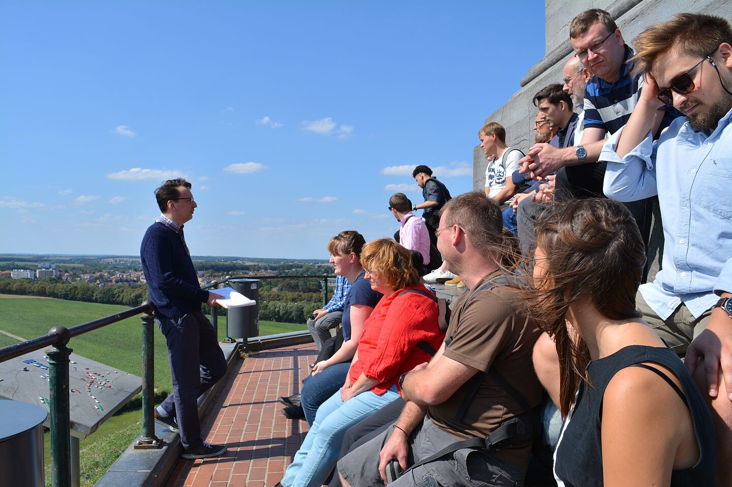 Mitglieder einer Reisegruppe sitzen in erhöhter Position an einem Aussichtspunkt. Der Himmel ist beinahe wolkenlos, ein Dozent steht der Gruppe zugeneigt am Geländer vor ihnen.