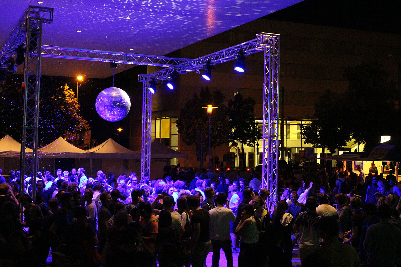 Tanzfläche bei Nacht vor der Bühne mit großer Diskokugeln und vielen Tanzenden