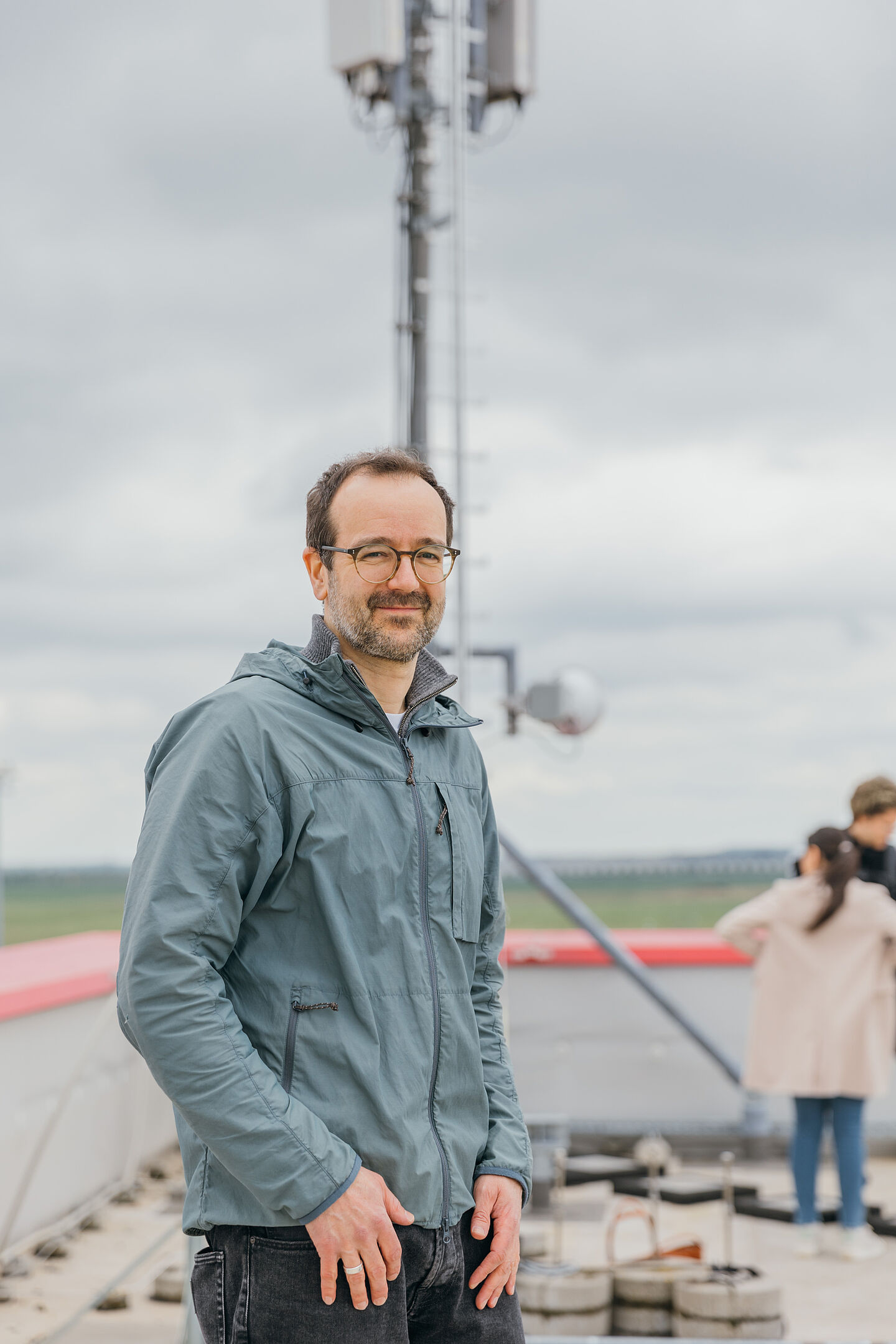 Lächelnde Person in Windjacke steht auf einem Dach vor einem Mast.