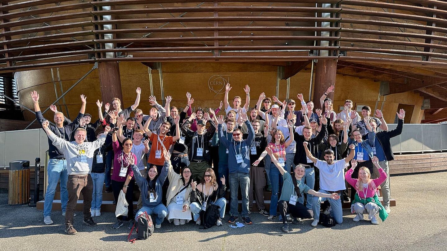 Eine Gruppe freudig winkender junger Menschen vor dem Haupteingang des CERN.