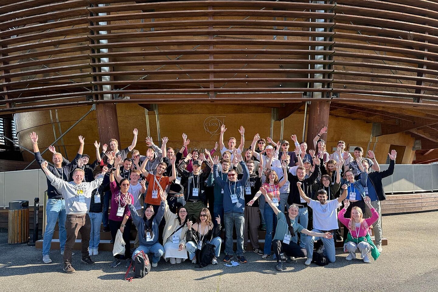 Eine Gruppe freudig winkender junger Menschen vor dem Haupteingang des CERN.