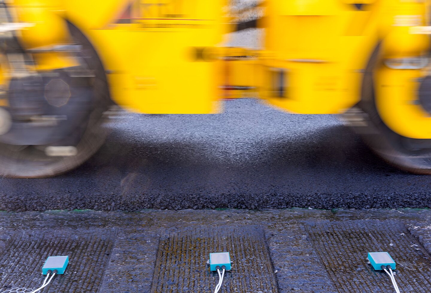 Im Hintergrund verdichet ein Walzenfahrzeug die Straße mit den frisch asphalttierten Sensoren