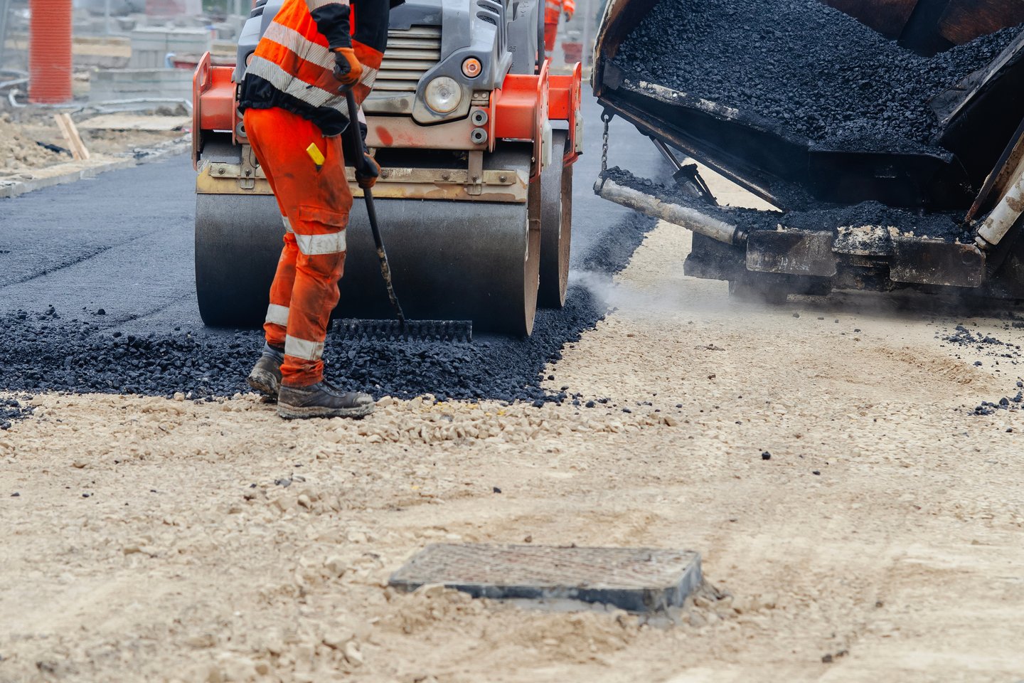Mann arbeitet im Straßenbau, hinten fährt Walze über Straße