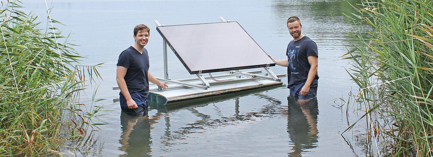Zwei Männer stehen im Wasser eines Sees und halten stolz eine schwimmende Plattform mit einem Solarpanel.