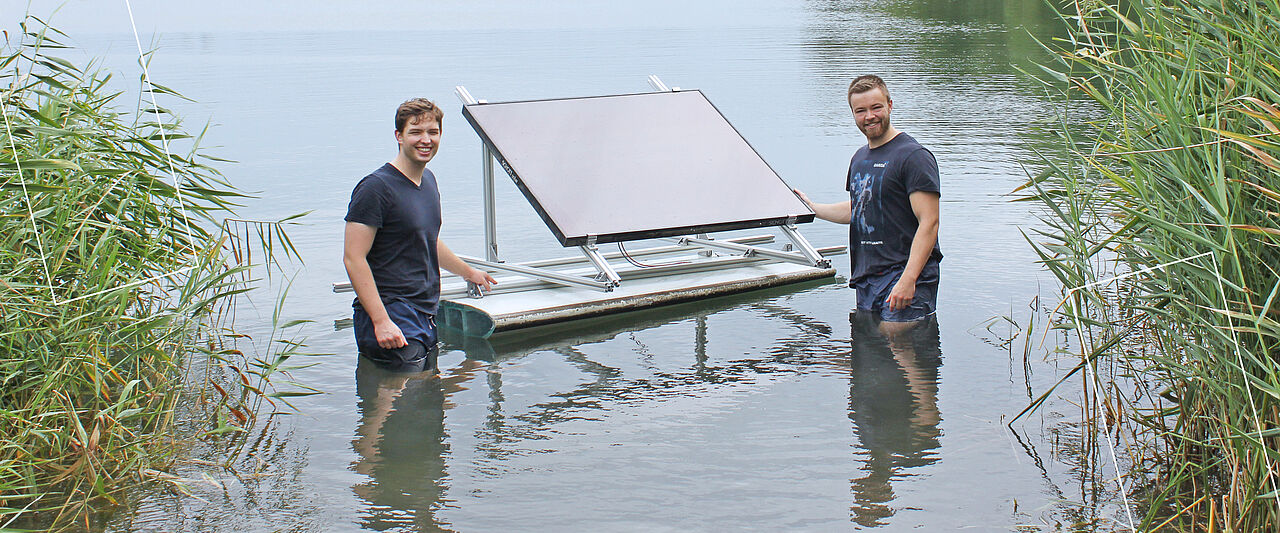 Zwei Männer stehen im Wasser eines Sees und halten stolz eine schwimmende Plattform mit einem Solarpanel.