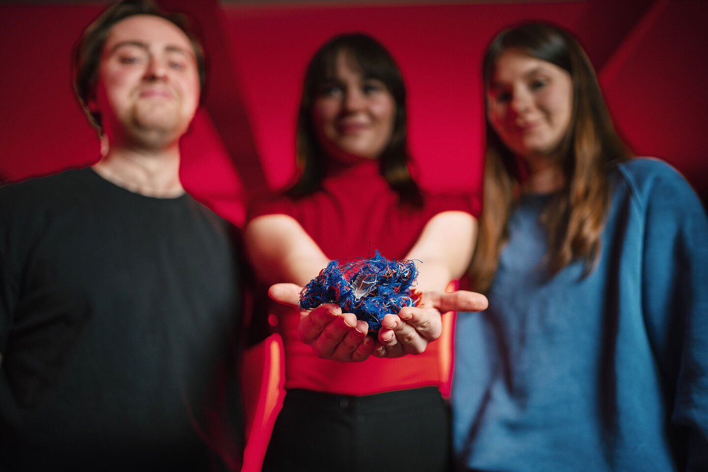 Das in rotes Licht getauchte Bild zeigt das Fiber Flow Team mit von links nach rechts: Verfahrenstechniker Ludwig Morgenstern, Umweltingenieurin Lena Strobl und Wirtschaftsingenieurin Elena Richter. Alle drei Personen sind unscharf bis auf die Hände von Lena Strobl, in denen sie textilen Abfall hält.