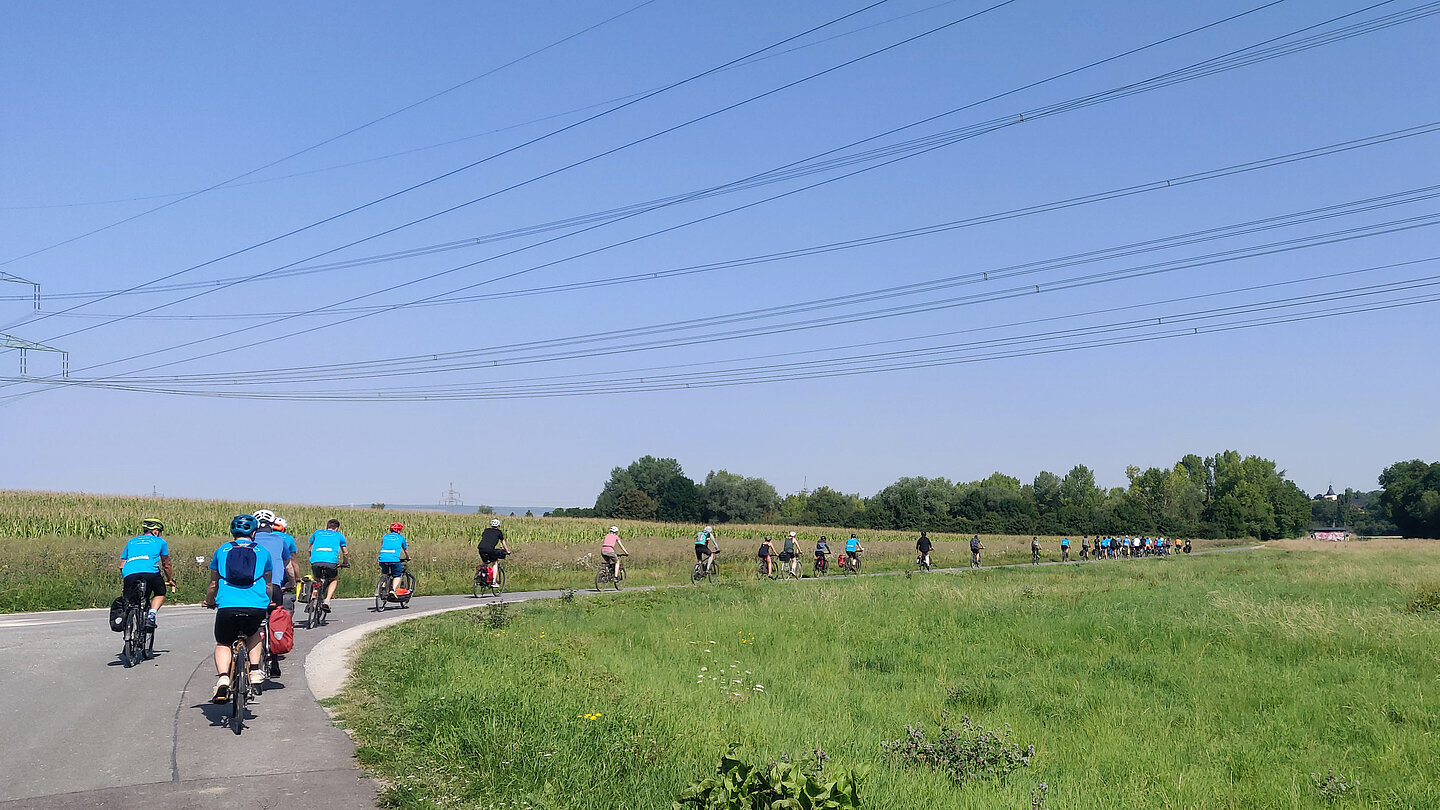 Ein Gruppe Menschen radelt auf einer Straße durch Felder.