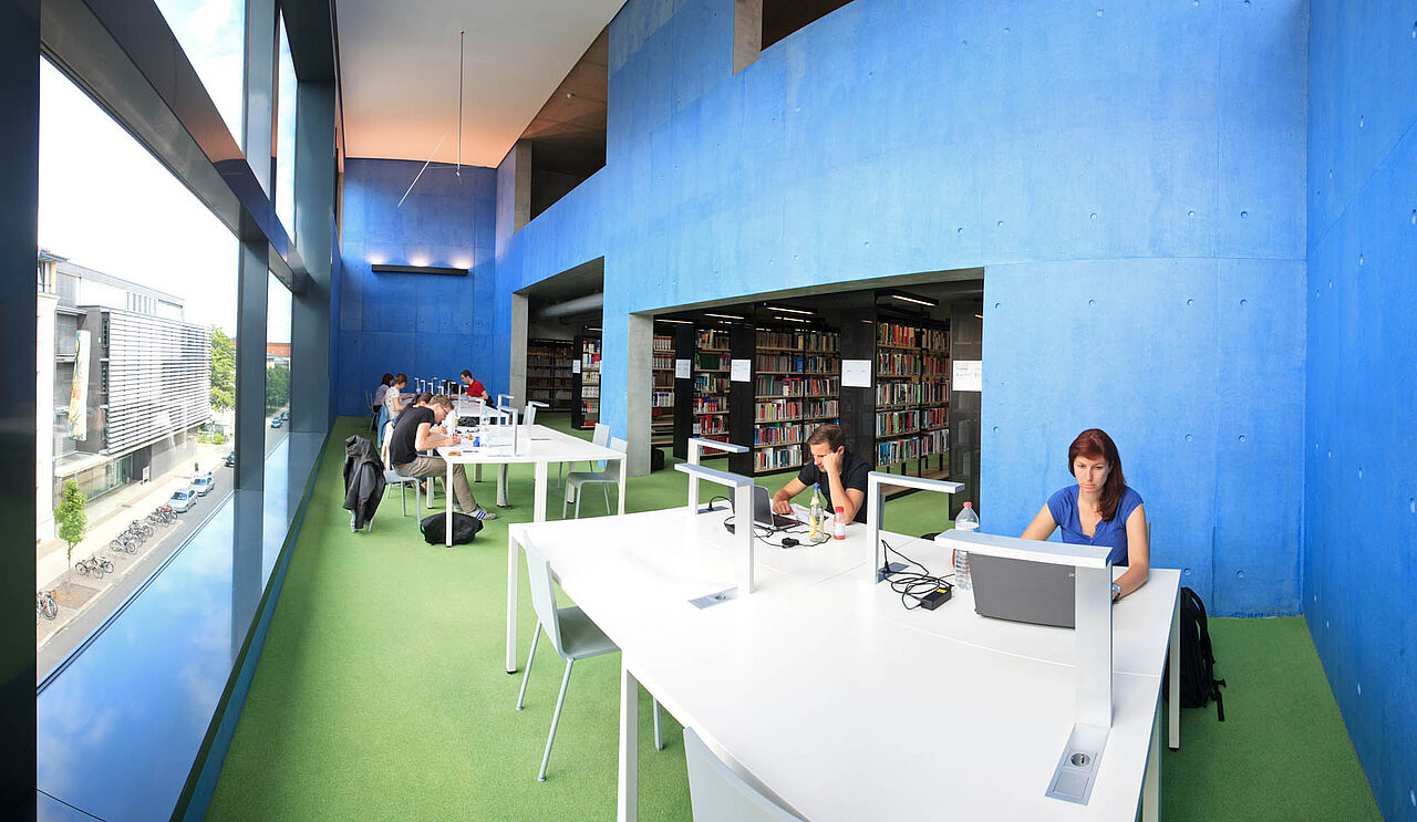 Bibliothek Innenansicht. Der Raum ist geräumig, es fällt viel Licht von einer Fensterwand ein. Die Wände sind in blau gestrichen. In der Mitte stehen mehrere weiße Tische an denen Menschen arbeiten. 
