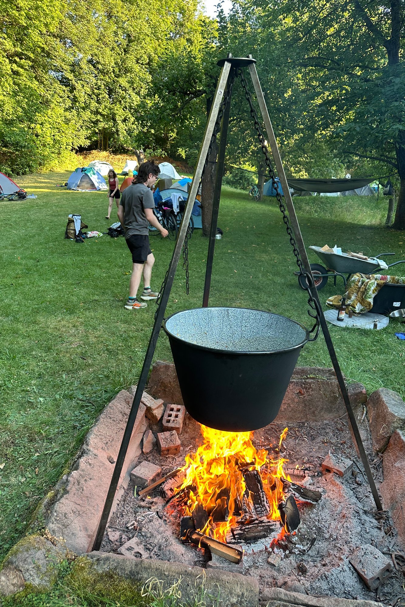 Eine Feuerstelle mit Dreibein und Kochkessel auf einem Campingplatz mit Zelten.
