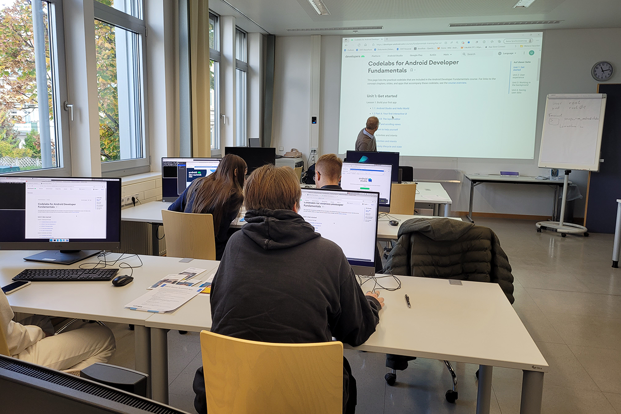 Schülerinnen und Schüler sitzen in einem Seminarraum an Computern und entwickeln ihre eigene App.
