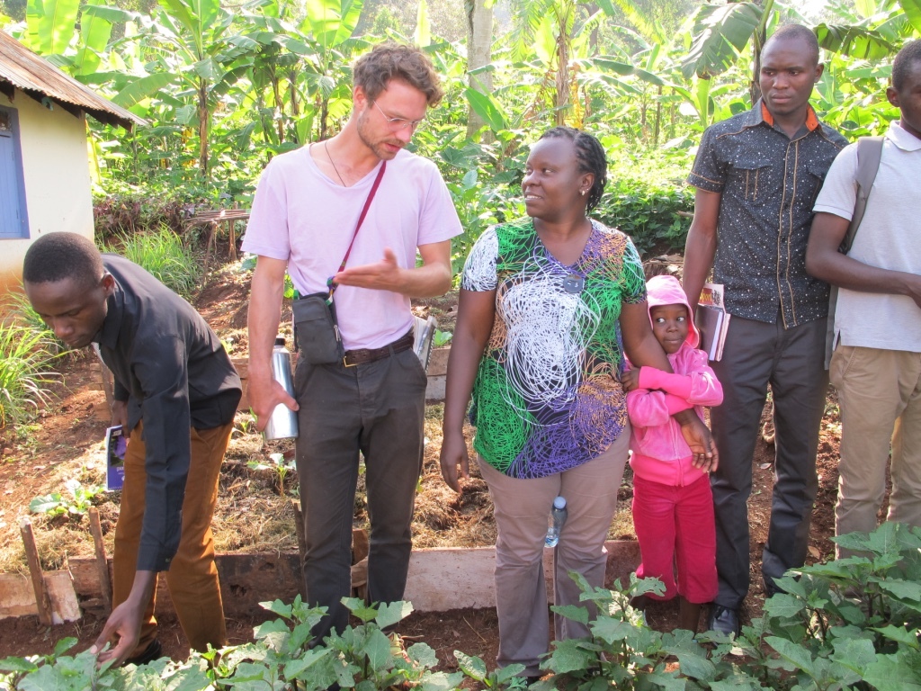 Ruben Gerstner unterhält sich an einem Beet mit einer Dozentin über das studentische Gartenbauprojekt. Die Dozentin hält ein Kind am Arm, weitere junge Männer inspizieren das Beet und haben Schreibmaterialien in der Hand.