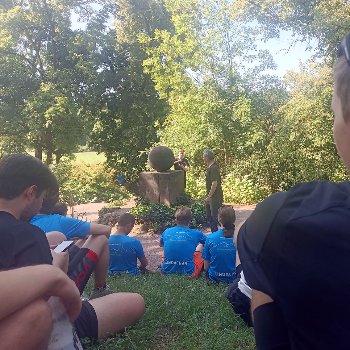 Eine Gruppe Menschen sitzt auf einer Wiese vor einem Denkmal, einer auf einem Würfel ruhende Kugel aus Sandstein..