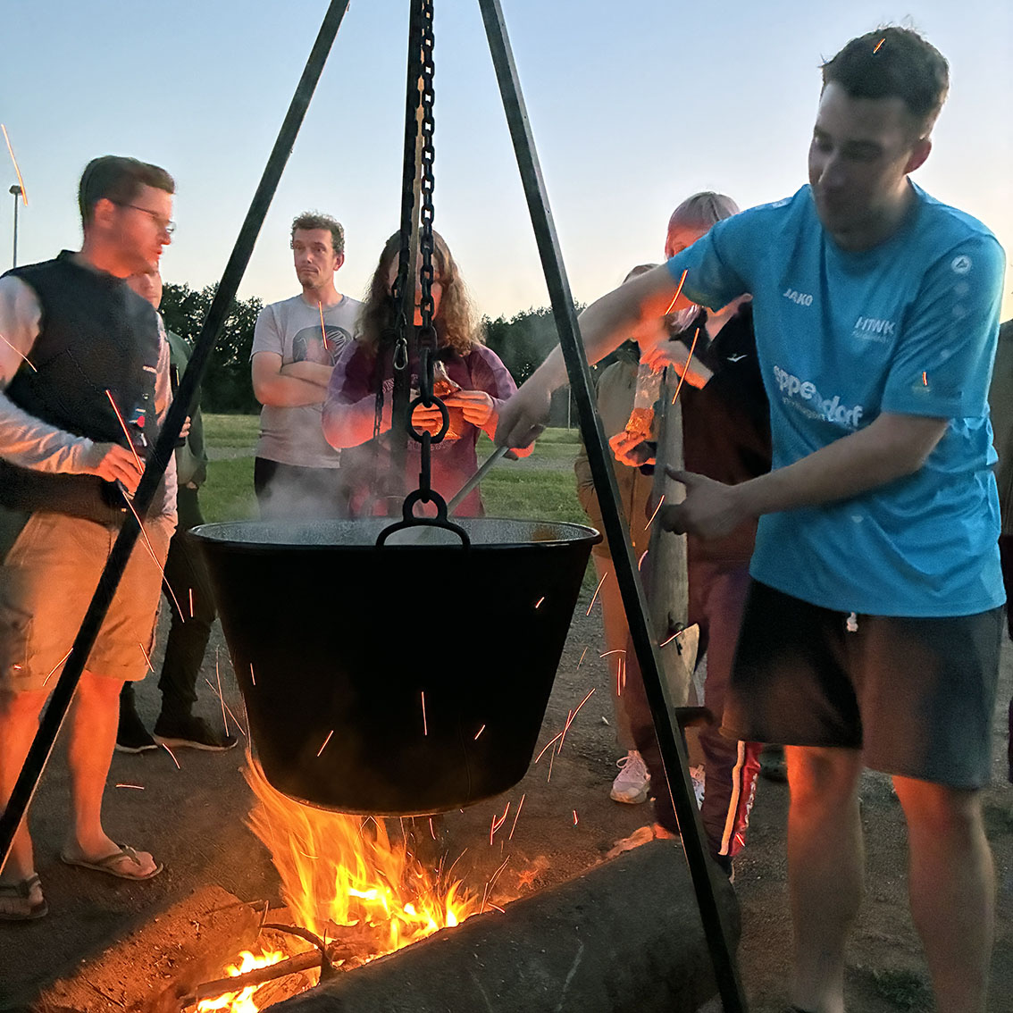 Ein Kessel hängt über einem Lagerfeuer. Mehrere Personen stehen ringsherum, einer rührt im Kessel.