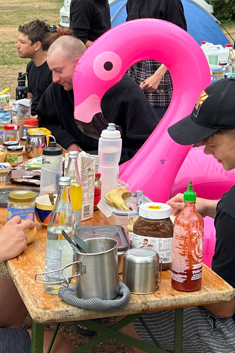 Mehrere Personen an einem üppigen Frühstückstisch unter freiem Himmel, zwischen ihnen sitzt ein aufblasbarer Flamingo-Schwimmreif auf der Bank.