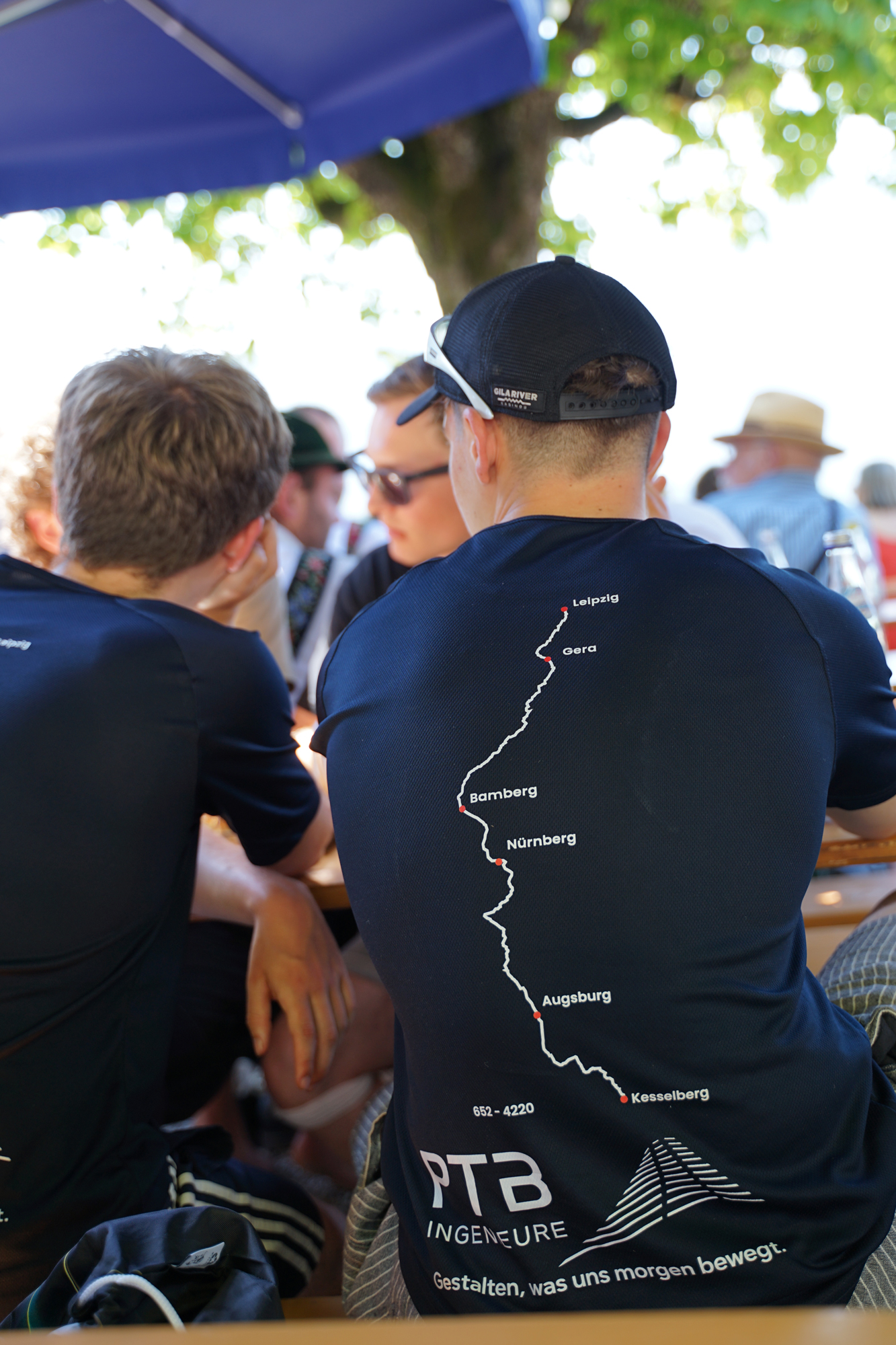 Mehrere Menschen sitzen in einem Freisitz. Eine Person im Fokus trägt das gemeinsame Teamshirt, auf dem die Radroute der Tour angebildet ist.
