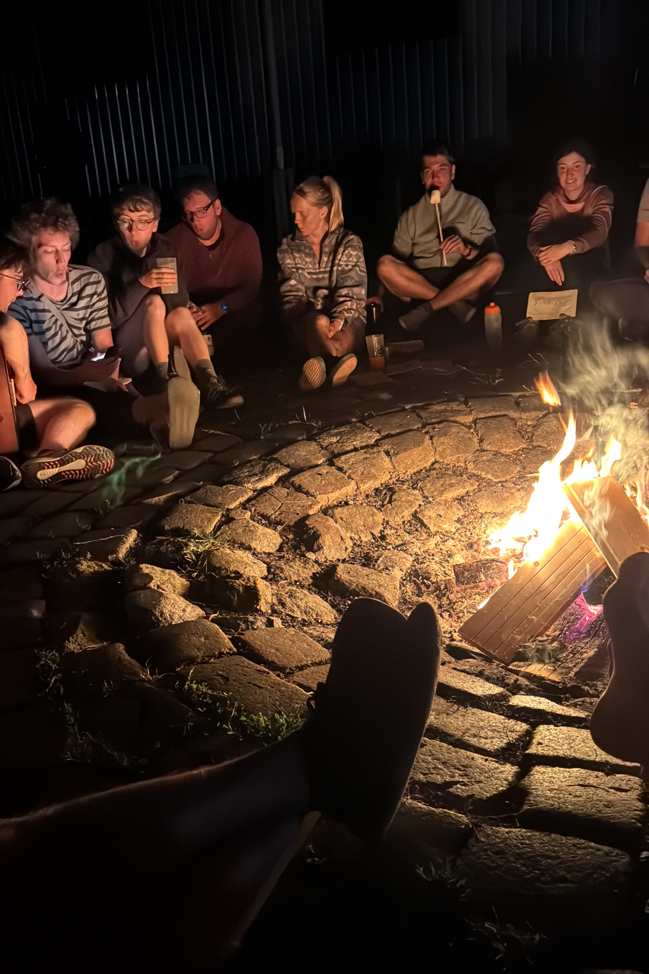 Mehrere Menschen sitzen um ein großes Lagerfeuer bei Dunkelheit und singen gemeinsam.