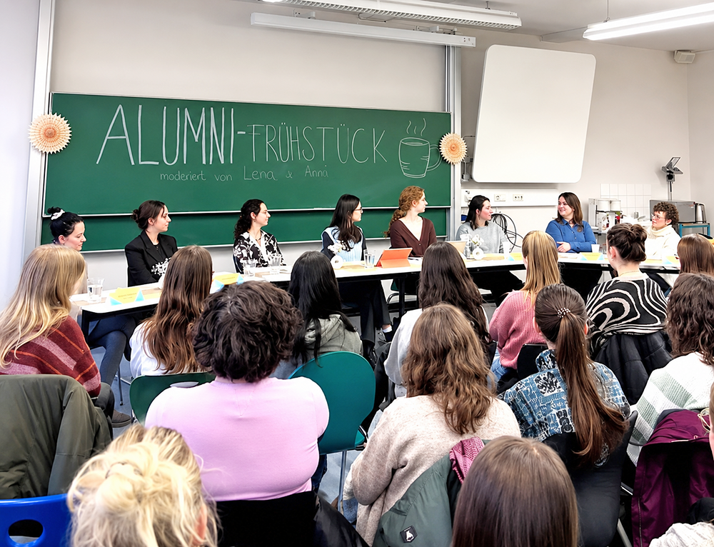 8 Almunae sitzen vor einer Tafel im Alumni-Frühstück 2026 vor einer studentischen Hörerschaft