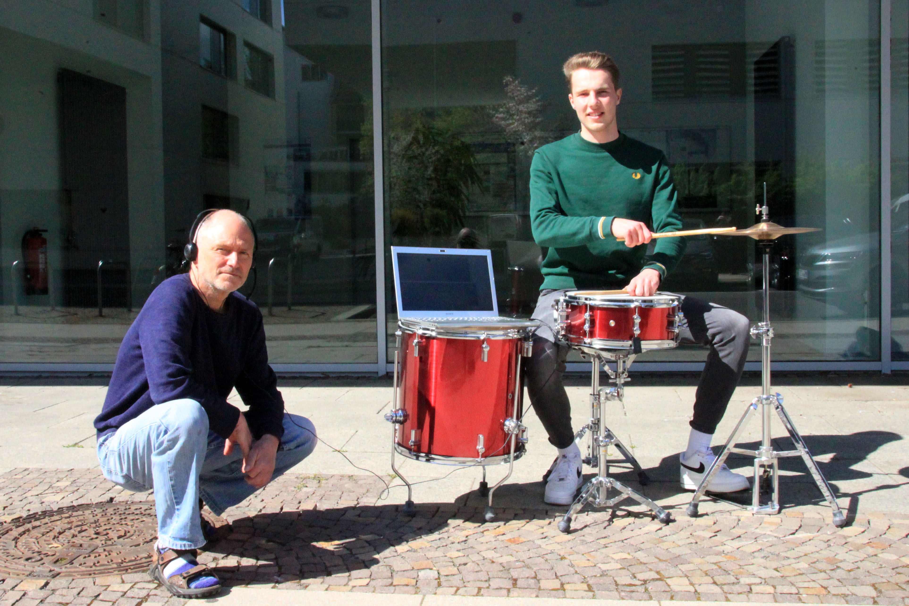 In der Sonne steht ein rotes Schlagzeug, daran sitzt Florian Wedhorn und hält die Schlagzeugstöcke auf die Trommeln; auf einer der Trommeln steht ein Laptop und daneben hockt Professor Christian Weickhardt mit Kopfhörern
